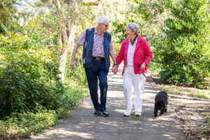 Jim and Julie walking hand in hand with a dog along a leafy garden path at Sundale Coolum Waters Retirement Village.